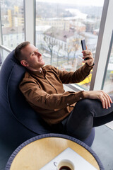 Young successful male businessman is sitting near a large window in the office and relaxing. Rest after a working day