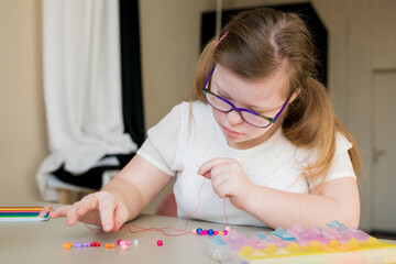 Cute teen girl stringing beads and making bracelet. Down syndrome kid crafting jewelry and...