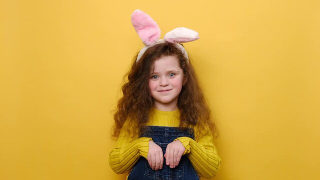 Cute little girl child keeps hands as if rabbit, wears soft bunny ears, has friendly look, smiles cheerfully, ready to congratulate children with coming Easter, models on yellow wall. I am lovely hare