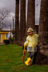 a little girl, wearing a raincoat with a hood and an umbrella in her hand.standing near a tree