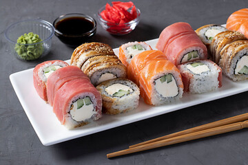 Sushi set with salmon, tuna and smoked eel with philadelphia cheese on white plate on dark gray background. Served with soy sauce, wasabi, pickled ginger and sticks for sushi.