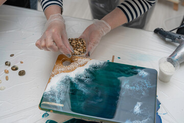 artist's hands decorating a wooden serving board with seashells and stones, epoxy resin, selective...