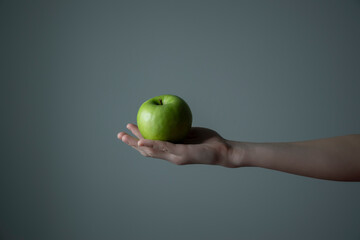 Woman`s hand holding green apple grey neutral background - raw photo. Dietary food and vitamins concept.  Non-filtered template for product advertising. 