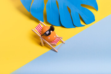 Creative funny composition with Easter egg with sunglasses while sitting on deck chair on illuminating yellow and pastel blue background