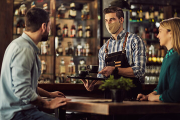 Happy waiter with face mask serving coffee to a couple in a cafe,