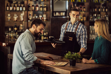 Happy waiter wearing protective face mask while serving coffee to customers in a cafe.