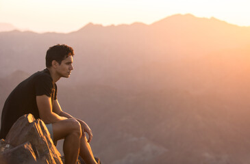 Attractive young man sitting alone relaxing and enjoying the view on top of a mountain peak at beautiful sunrise. 