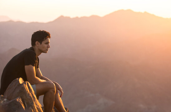Attractive young man sitting alone relaxing and enjoying the view on top of a mountain peak at beautiful sunrise. 
