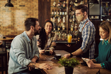 Happy waiter with face mask serving coffee to group of friends in a cafe.