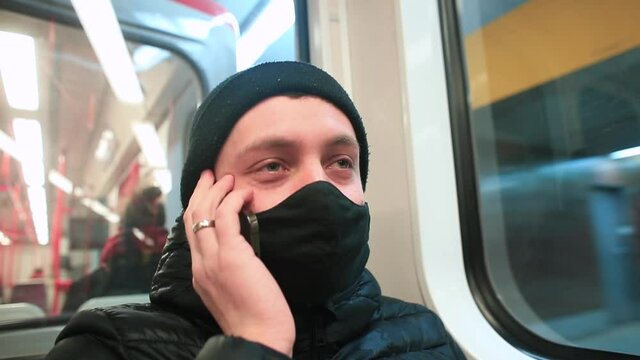 A masked male passenger talks on the phone in a subway car. He holds the gadget to his ear. Train movement at the metro station. Oral message transmission using telephony.