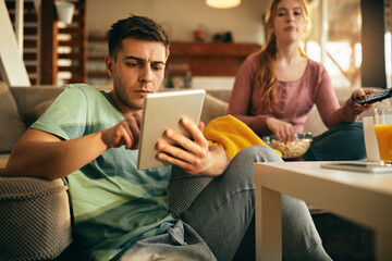 Young man surfing the net on touchpad while relaxing with his girlfriend at home.