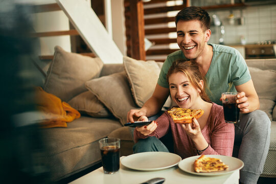 Happy Couple Watching Something Funny On TV While Eating Pizza At Home.