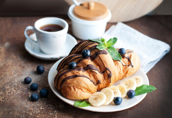 Croissant with chocolate, blueberries, bananas, and mint on a dark brown background. Breakfast with a croissant and a cup of coffee
