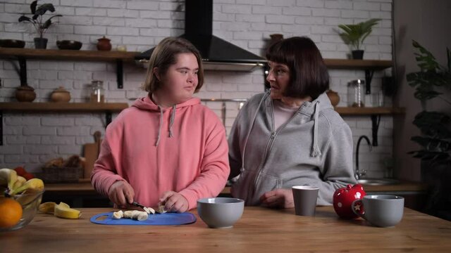 Loosing weight handicapped teenage girl with down syndrome preparing healthy fruit snack and talking with mother in kitchen. Overweight teen child and her mom spending leisure at home