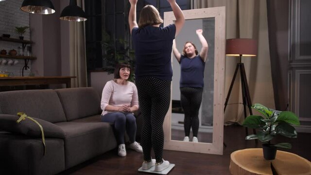 Slimming overweight teenage girl with down syndrome getting on scales in front of mirror. Happy handicapped teenager rejoicing at results of weight loss and hugging supporting her mother