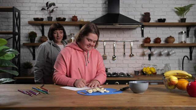 Curvy disabled teenager enjoying drawing with pencils while her mom doing household chores in home kitchen. Cheerful teen girl with down syndrome hiding drawing from curious mother