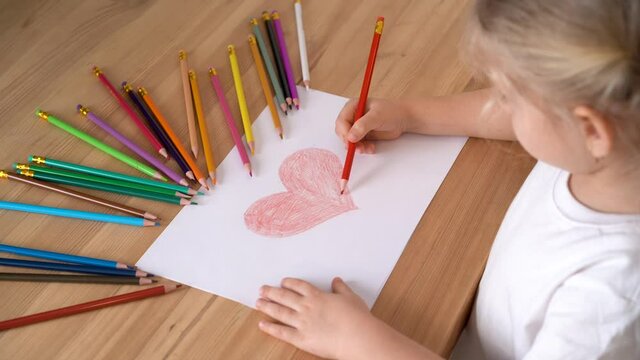 the hands of a small child draw a heart with a red pencil on a white piece of paper, lots of colored pencils around.