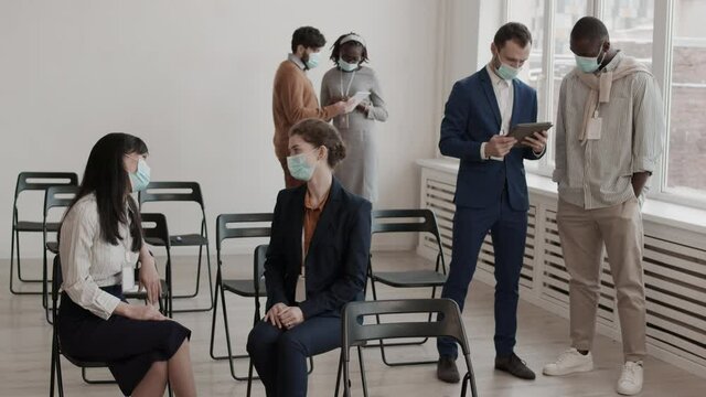 Steadicam medium long of Asian and Caucasian female colleagues sitting on chairs in conference room, chatting in foreground of diverse coworkers standing, people wearing medical face masks
