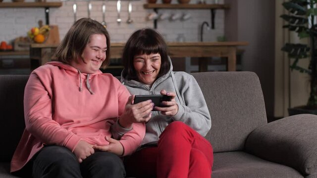 Close-up of cheerful disabled girl with down syndrome and her mom laughing while watching video on cellphone. Positive child with mother enjoying funny content on smartphone relaxing at home