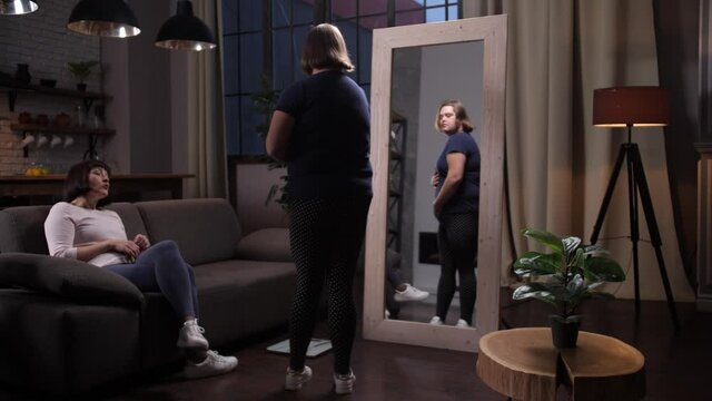 Curvy teen girl with down syndrome looking in large mirror critically examining her appearance after weight loss. Handicapped plus size child in tight clothes needing mom's support to lose weight
