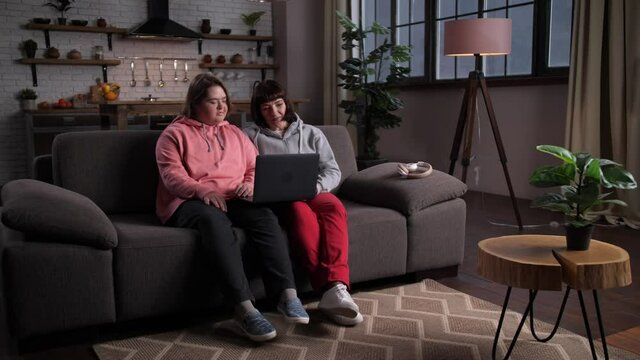 Young woman with down syndrome together with her mature mother waving hello while communicating via internet with relatives. Positive mom and handicapped daughter sitting on couch during online chat