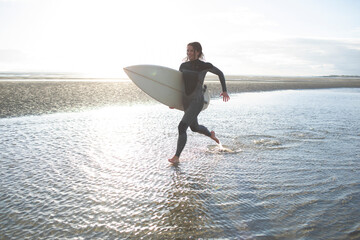 Young female surfer running with surfboard in sunny ocean beach surf