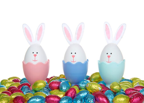 Row of Easter Eggs crafted into bunnies with felt ears, sitting in colorful egg cups with colorful foil wrapped chocolate egg candies, isolated on white background.