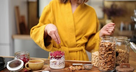 Young woman in yellow bathrobe making breakfast with a yogurt and raspberry, decorated meal in the glass bowl. Healthy vegan cereal meal for breakfast concept - Powered by Adobe