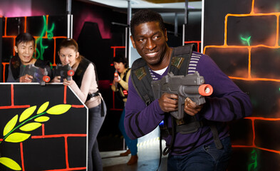 Fototapeta na wymiar Emotional African man with his laser pistol playing laser tag with friends at the dark labyrinth