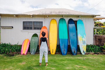 girl walking with surfboard in front of wall of longboards