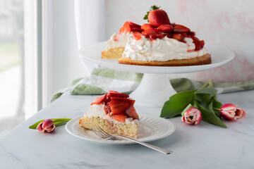 Close up view of a slice of strawberry shortcake against a sunny window with the full cake in behind.
