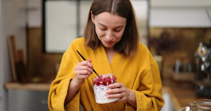 Hungry woman dressed in yellow bathrobe eats yummy cereal breakfast with yogurt and berries from the bowl on the kitchen at home