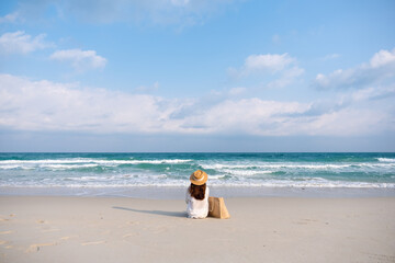 Rear view image of a woman with hat and bag sitting on the beach with blue sky background