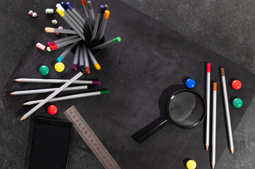 colored pencils in a glass and against the background of a chalkboard with colored magnets, a magnifying glass, a ruler and a notebook, top view, back to school