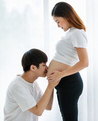 Fototapeta premium portrait of a very lovely young Asian couple. A husband sitting and kissing a wife's belly while a pregnant woman standing smiling and looking at a man with much love
