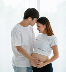 portrait of a young happy Asian couple standing together by the window at home. A husband kissing a pregnant wife's forehead with care and love while touching a lady's belly