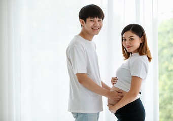 A lovely young Asian couple husband and wife standing together smiling and touching a pregnant woman's belly with love and care. Happy and healthy family concept.