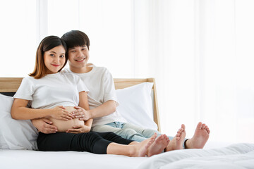 portrait of a good-looking young Asian man and woman wearing a white nightdress sitting on a bed together. They are smiling happily and touching the belly of a pregnant mother