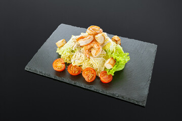 Caesar salad with shrimps. On a black slate board. Salad ingredients: cherry tomatoes, salad mix, croutons, parmesan cheese, sauce. Dark background. Isolated. Close-up.
