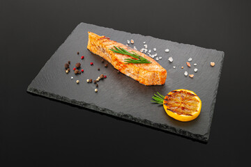 Grilled salmon fillet. On a black slate board. Decorated with pepper, sea salt and lemon wedge. Dark background. Isolated