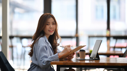 Young businesswoman holding clipboard and smiling to camera while sitting at modern office.