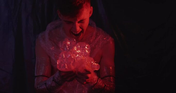 Plastic pollution. Ecology problem. Waste recycling. Frustrated pissed off man in polyethylene film yelling at creased used bottles in red neon light on dark background.