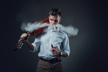 Elegant and also stylish man in suit poes in dark background smoking electric cigarette and holding acoustic guitar on his shoulder.