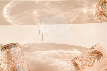 Still life scene with wine glasses on beige background in sunlight and blank business, greeting card, invitation mockup. Long harsh shadows.