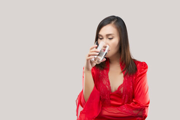 Sexy girl drinking a glass of water before bed on a gray background. Asian woman wearing red satin...