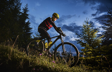 Naklejka premium Smiling young man in sports cycling suit riding bicycle downhill with beautiful blue sky on background. Male bicyclist wearing safety helmet and glasses while cycling down grassy hill at night.