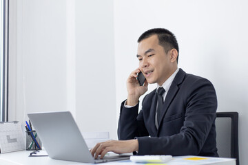Asian man employee talking on the phone and working in a laptop with a smiling face, Office staff work and telephone conversations, Business conversation over the phone, handsome male in a black suit.