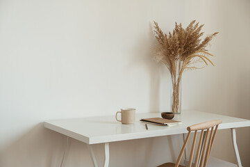 Women's home office workspace. Freelance work, business workplace. Light aesthetic hygge space with wooden chair, table, reed pampas grass bouquet, mug, notebook. Modern interior design.