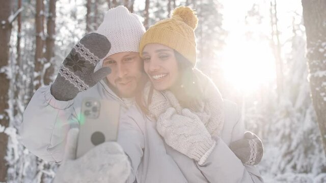 Young couple walks in a winter forest, man and female take a selfie on a smartphone against the background of a winter forest, they make faces at the camera, sunny winter weather.