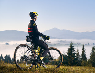 Back view of man in cycling suit standing with bicycle on grassy hill and looking at beautiful misty mountains. Male bicyclist in safety helmet enjoying view of majestic mountains during bicycle ride.
